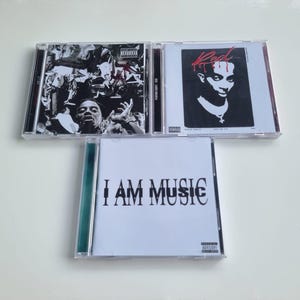 May include: Three CD cases. The top left case features a monochrome image of a crowd. The top right case has a black and white portrait with the word "Red" in red. The bottom case reads "I AM MUSIC" in black.