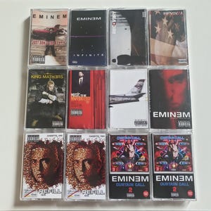 Puede incluir: Una colección de doce cintas de casete con portadas de álbumes de Eminem. Las cintas muestran varios títulos de álbumes y portadas, incluyendo imágenes del artista, banderas y otros gráficos. Las cintas están en estuches de plástico transparente.