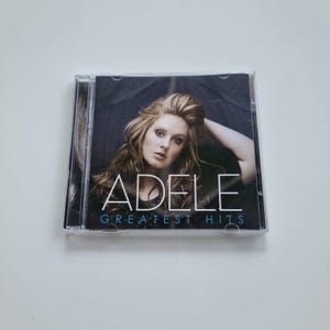 Può includere: Un'immagine di un CD con l'immagine di Adele e il testo "ADELE GREATEST HITS" in bianco e blu. La copertina dell'album mostra Adele con lunghi capelli castani ondulati, su sfondo scuro. La custodia del CD è in plastica trasparente.