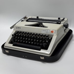 May include: A white Olympia Super SM 1 typewriter with a black keyboard and a black carrying case. The typewriter has a red power switch and a black return lever. The carrying case has two silver clasps.