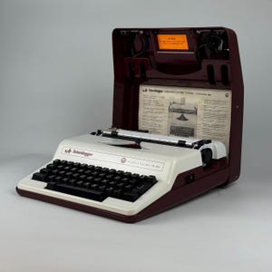 May include: A white ceramic mug with a black handle and the text "But First, Coffee" in a bold, black font. The mug is set against a blurred background, suggesting a cozy setting.