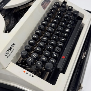 May include: A white Olympia Regina de Luxe typewriter with a black case. The typewriter has a black keyboard with white lettering and a red dot on the return lever.
