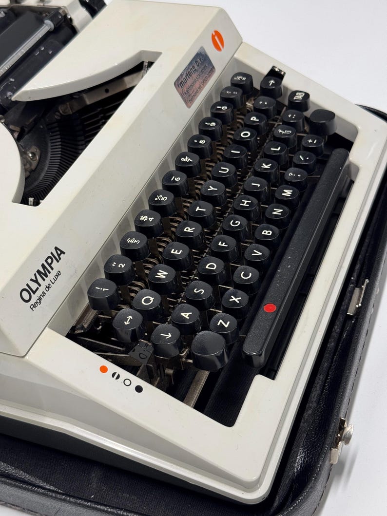 May include: A white Olympia Regina de Luxe typewriter with a black case. The typewriter has a black keyboard with white lettering and a red dot on the return lever.