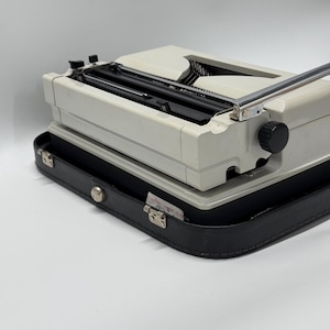 May include: A white vintage typewriter with a black carrying case. The typewriter has a silver carriage return lever and a black knob on the right side.