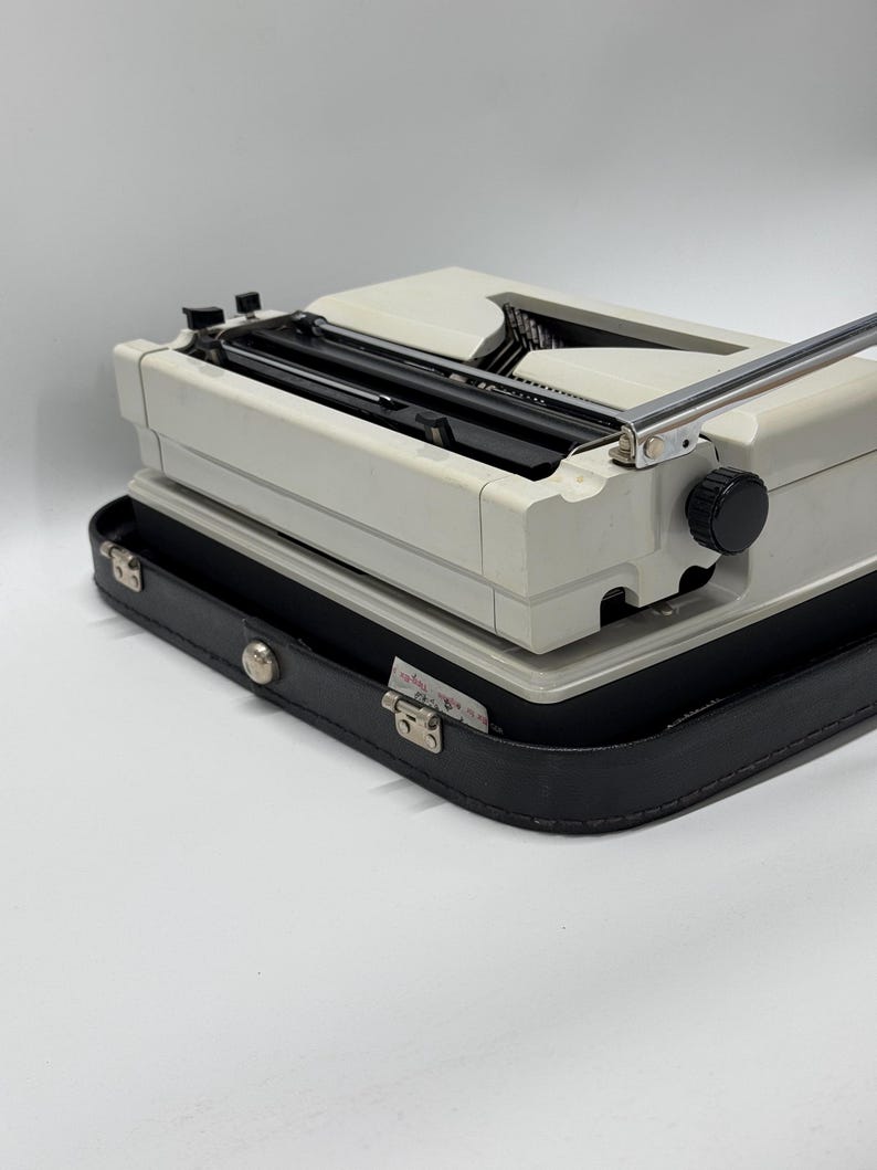 May include: A white vintage typewriter with a black carrying case. The typewriter has a silver carriage return lever and a black knob on the right side.