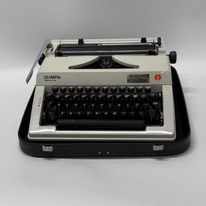 May include: A white Olympia typewriter with a black case. The typewriter has a black keyboard with white keys and a black carriage return lever. The typewriter is in a black carrying case with a silver latch.