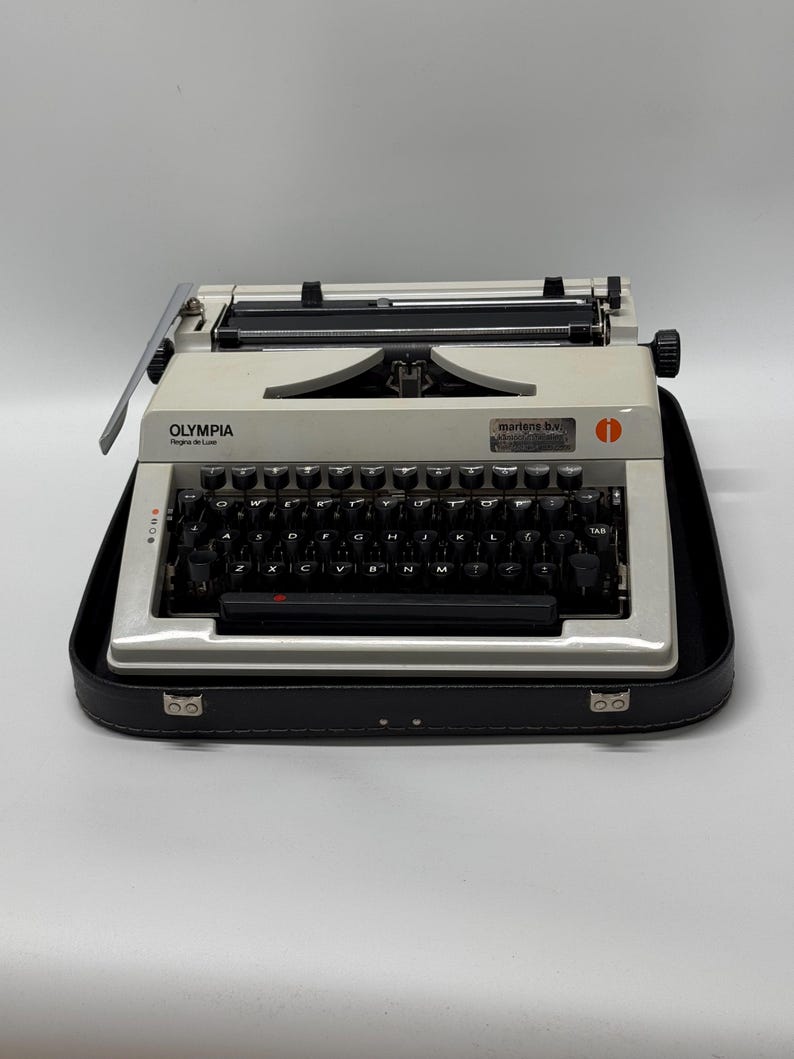 May include: A white Olympia typewriter with a black case. The typewriter has a black keyboard with white keys and a black carriage return lever. The typewriter is in a black carrying case with a silver latch.