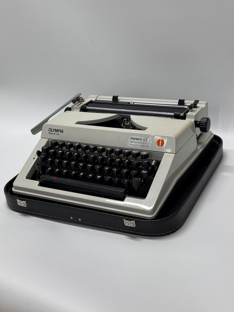 May include: A white Olympia Super SM 1 typewriter with a black keyboard and a black carrying case. The typewriter has a red power switch and a black return lever. The carrying case has two silver clasps.