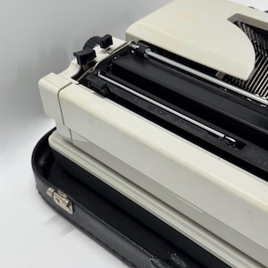 May include: A white vintage typewriter with a black case. The typewriter has a black carriage return lever and a black ribbon spool.