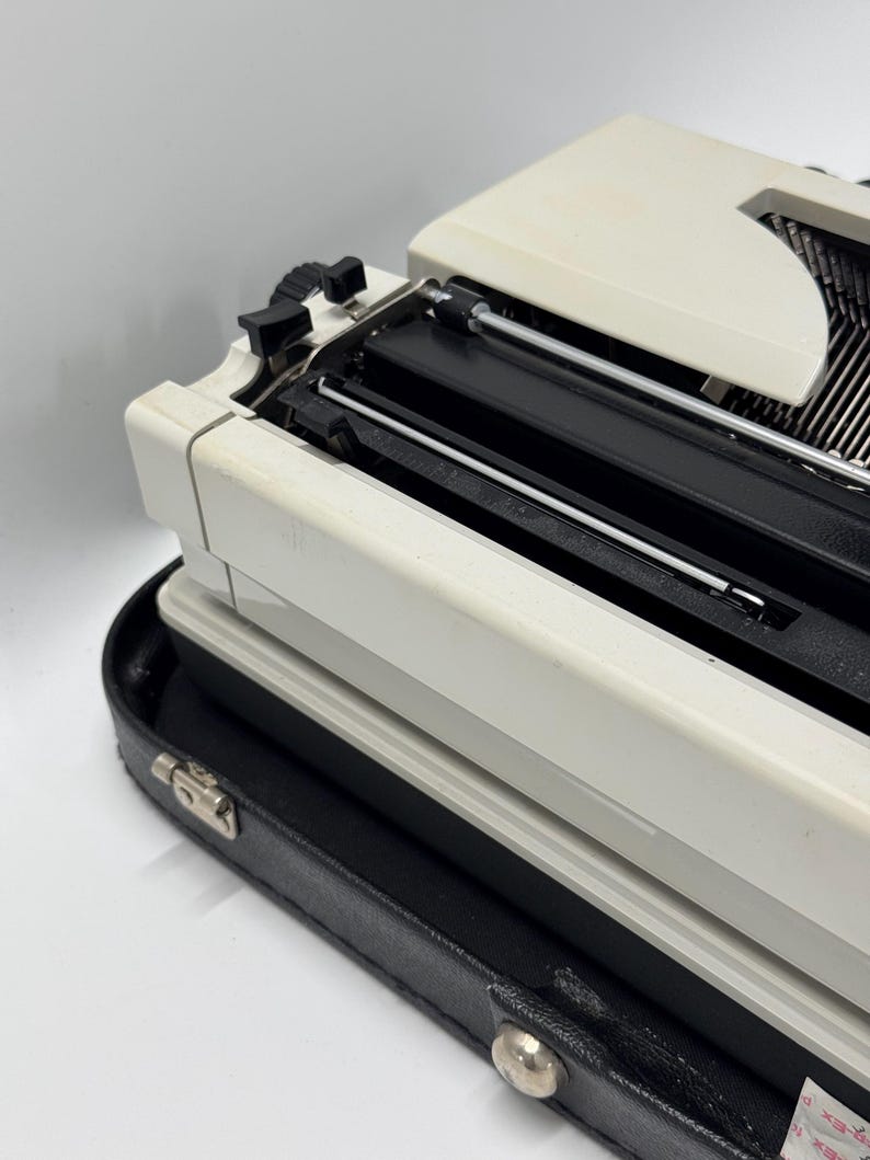 May include: A white vintage typewriter with a black case. The typewriter has a black carriage return lever and a black ribbon spool.