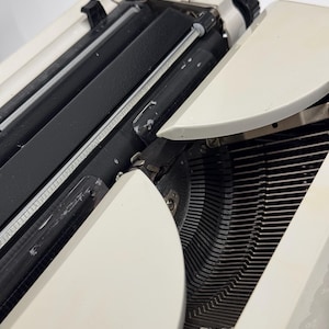 May include: A white vintage typewriter with a black carriage return lever and black keys. The typewriter is open and the type bars are visible.