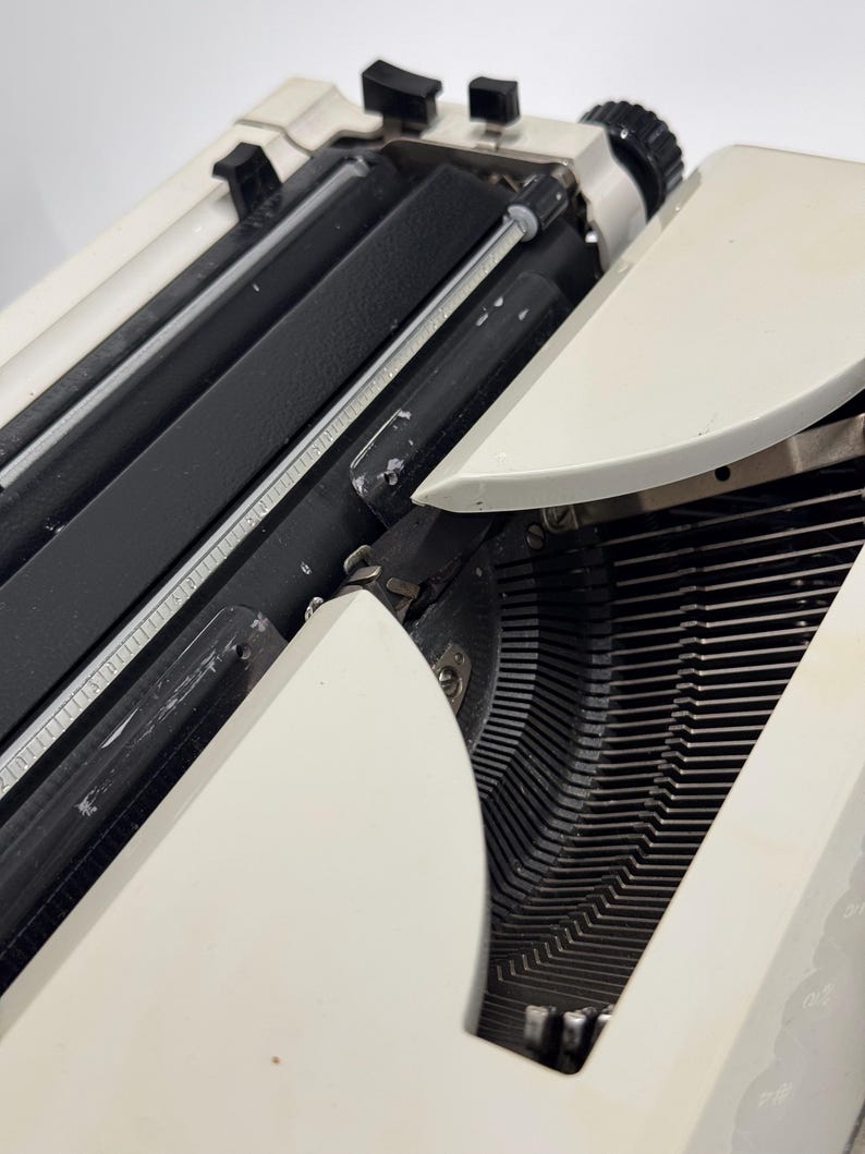 May include: A white vintage typewriter with a black carriage return lever and black keys. The typewriter is open and the type bars are visible.