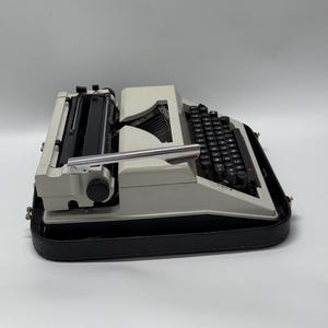 May include: A white vintage typewriter with a black keyboard and a black carrying case. The typewriter has a silver carriage return lever and a black knob on the side.