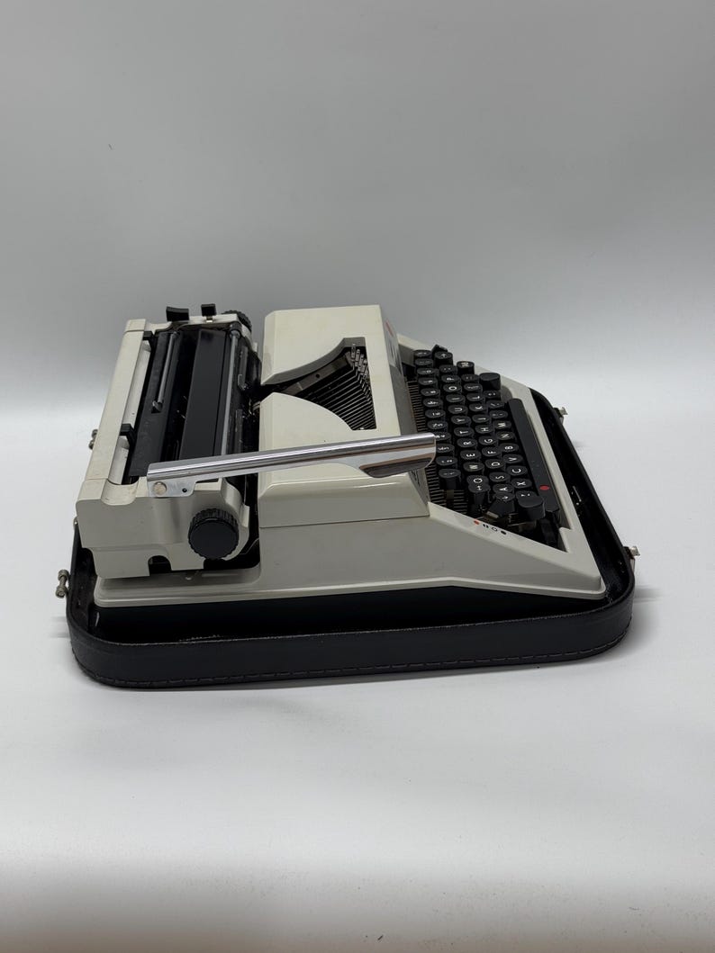 May include: A white vintage typewriter with a black keyboard and a black carrying case. The typewriter has a silver carriage return lever and a black knob on the side.