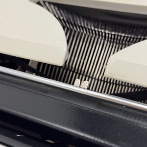 May include: Close-up of a vintage typewriter with a white plastic body and black metal carriage. The carriage is open, revealing the metal type bars and the paper roller.