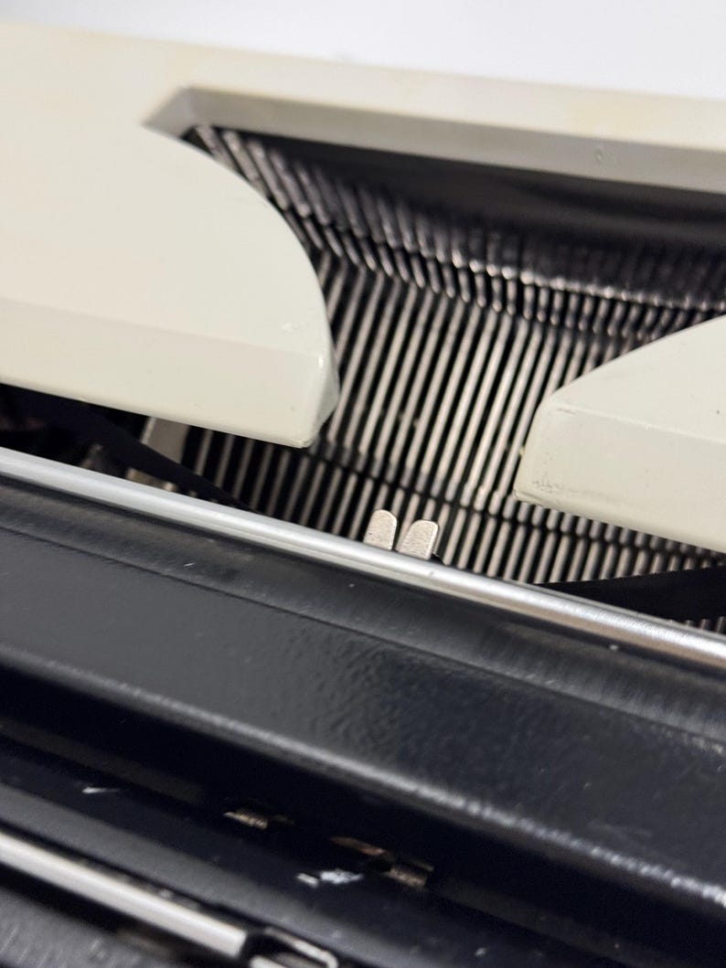 May include: Close-up of a vintage typewriter with a white plastic body and black metal carriage. The carriage is open, revealing the metal type bars and the paper roller.