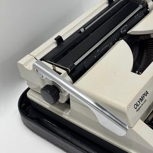 May include: A white Olympia Regina de Luxe typewriter with a silver carriage return lever and a black carrying case.