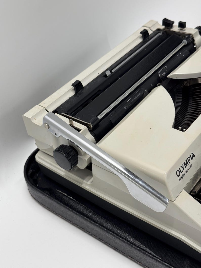 May include: A white Olympia Regina de Luxe typewriter with a silver carriage return lever and a black carrying case.