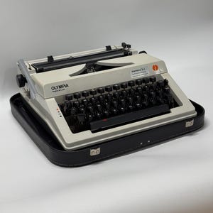 May include: A white Olympia Regina de Luxe typewriter with a black case. The typewriter has a black keyboard and a red sticker that says "martens b.v. Telefoonnummer 010-4112222".