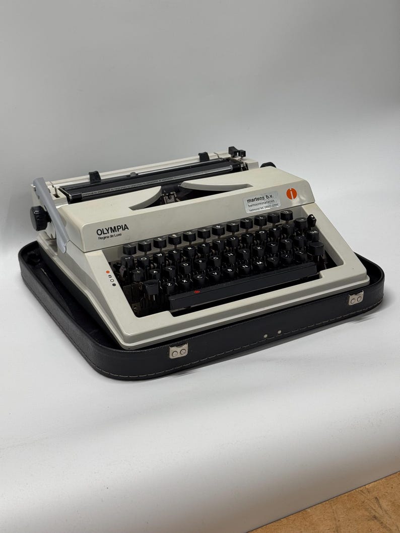 May include: A white Olympia Regina de Luxe typewriter with a black case. The typewriter has a black keyboard and a red sticker that says "martens b.v. Telefoonnummer 010-4112222".
