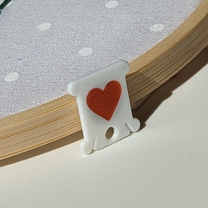 May include: A close-up of an embroidery hoop with light blue fabric featuring white polka dots. A white thread holder with a red heart is in the foreground. Dark green thread forms a stitched design on the fabric.