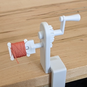 May include: A white thread winder with a clamp base, a gear, and a handle. The winder holds a spool of orange thread. The device is designed to be clamped to a table or other surface for use in sewing or crafting projects.