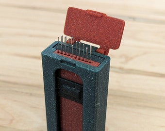May include: A dark gray, textured rectangular case with a hinged lid, containing a row of thin metal needles. The interior insert is a contrasting reddish-brown color, and a small black sliding mechanism is visible on the front of the case.