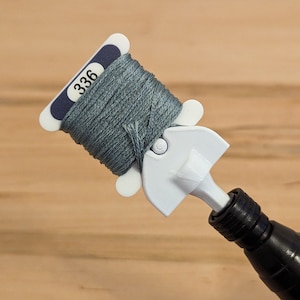Op de afbeelding: Een spoel teal borduurgaren, gemarkeerd met nummer 336, wordt vastgehouden door een wit en zwart gereedschap. De draad is strak opgerold, klaar voor borduren en andere handwerkprojecten.