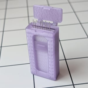 May include: A lavender-colored, rectangular case with a hinged lid, holding several silver needles. The case has a textured surface and a rectangular window. The lid is open, revealing the needles. The case is on a white surface.