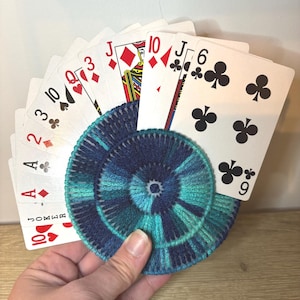 May include: A hand holding a fan of playing cards, including the Ace of Diamonds, 2 of Diamonds, 3 of Hearts, Queen of Hearts, and Jack of Diamonds. A blue and teal coaster is in the foreground.
