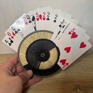 May include: A hand of playing cards fanned out, displaying various suits and numbers. The cards are held over a circular, embroidered coaster with a spiral design in black, gray, and beige. The cards include a Joker, 5 of hearts, and other cards.