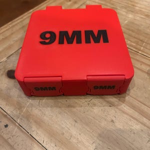 K&ouml;nnte beinhalten: Ein roter rechteckiger Koffer mit dem Text "9MM" in Schwarz auf der Oberseite und den Seiten. Der Koffer hat einen Klappdeckel und steht auf einer Holzoberfl&auml;che.