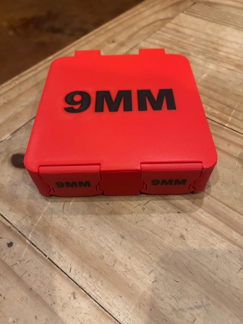 K&ouml;nnte beinhalten: Ein roter rechteckiger Koffer mit dem Text "9MM" in Schwarz auf der Oberseite und den Seiten. Der Koffer hat einen Klappdeckel und steht auf einer Holzoberfl&auml;che.