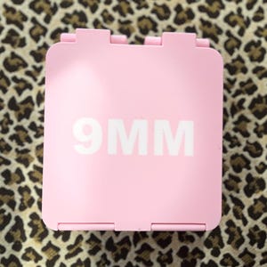 K&ouml;nnte beinhalten: Ein rosa quadratischer Beh&auml;lter mit dem wei&szlig;en Text "9MM" vor einem Leopardenmuster-Hintergrund. Der Beh&auml;lter hat ein Scharnierdesign und scheint aus Kunststoff zu sein.
