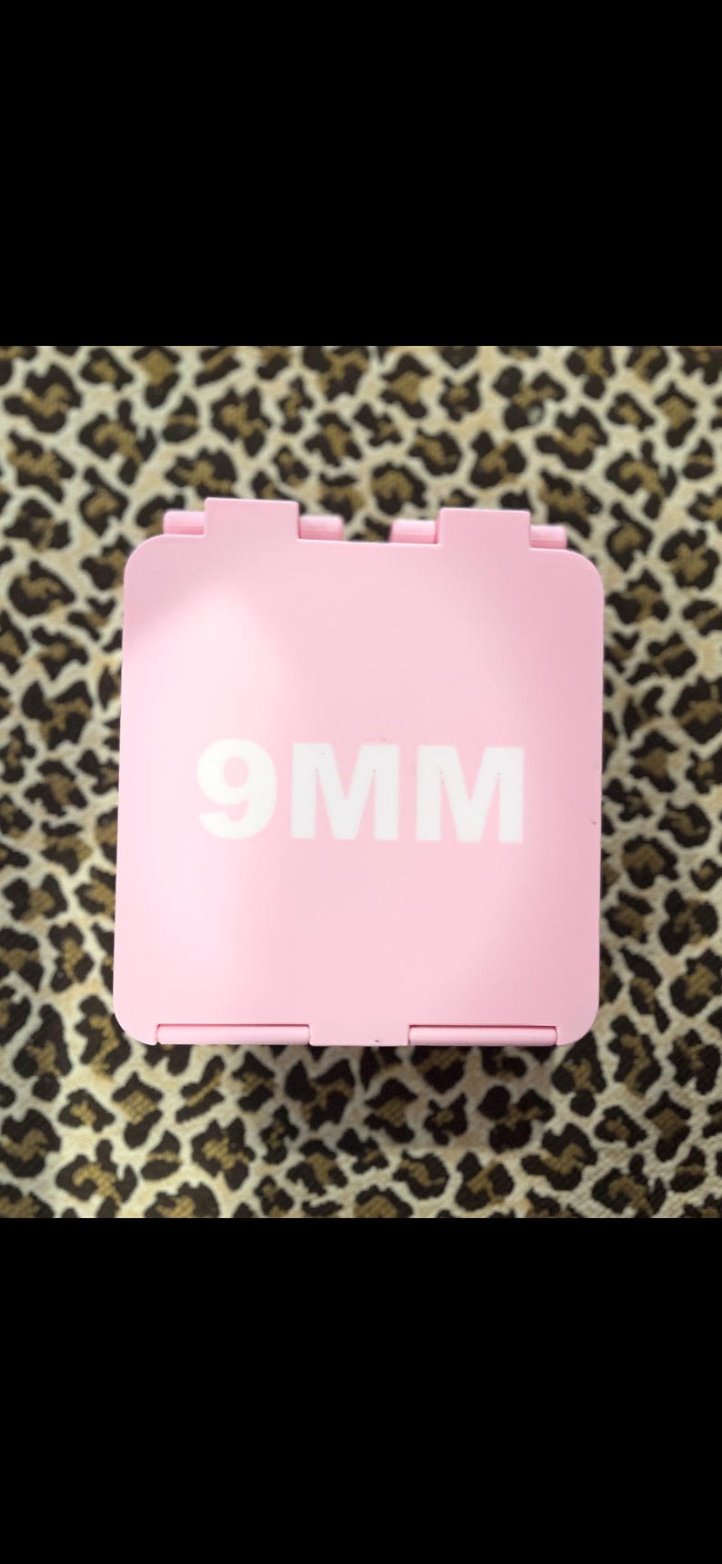 K&ouml;nnte beinhalten: Ein rosa quadratischer Beh&auml;lter mit dem wei&szlig;en Text "9MM" vor einem Leopardenmuster-Hintergrund. Der Beh&auml;lter hat ein Scharnierdesign und scheint aus Kunststoff zu sein.