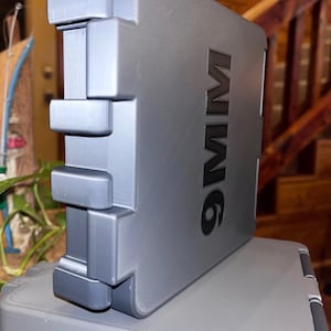 K&ouml;nnte beinhalten: Ein grauer, rechteckiger Koffer mit dem Text "9MM" auf der Seite. Der Koffer hat ein einzigartiges, geschichtetes Design mit hervorstehenden rechteckigen Formen an der Seite. Der Koffer steht auf einer grauen Oberfl&auml;che.