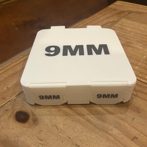 K&ouml;nnte beinhalten: Ein wei&szlig;er Kunststoffbeh&auml;lter mit dem Text "9MM" in gro&szlig;en, schwarzen Gro&szlig;buchstaben auf der Oberseite. Der Beh&auml;lter hat auch "9MM" auf der Vorderseite aufgedruckt. Der Beh&auml;lter steht auf einer Holzoberfl&auml;che.
