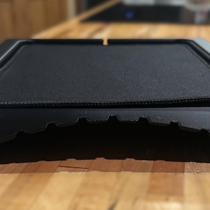 May include: A black, low-profile stand featuring a rectangular, textured top surface and a curved, notched base. The structure is constructed from a matte material, providing a stable platform with a distinct, arched front edge and a flat top.