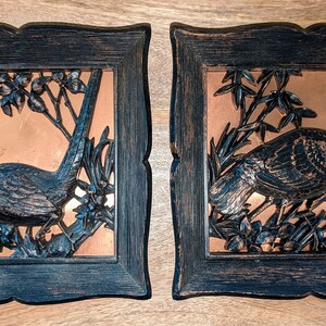 May include: Two framed wall art pieces. Each features a copper-colored square with a detailed relief of a bird perched on a branch. The frames are dark blue with a distressed finish, adding a vintage aesthetic.