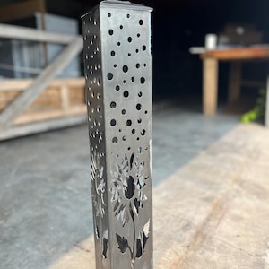 May include: A tall, rectangular metal garden torch with a sunflower design cut into the metal. The torch has a series of circular cutouts and a small, flat top. The metal has a silver finish.