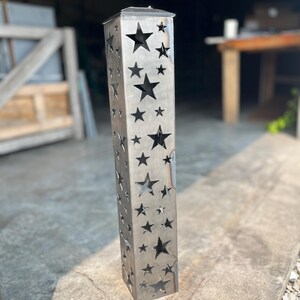 May include: A tall, rectangular metal lantern with a silver finish. The lantern features a series of star cutouts of varying sizes across its surface. The top has a small, flat lid. The lantern is designed to be placed outdoors.
