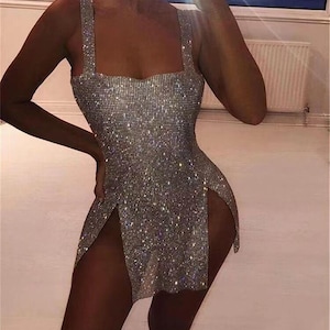 May include: A silver, sparkling mini dress with a square neckline and wide straps. The dress features high slits on both sides, creating a glamorous look. The dress is covered in shimmering embellishments.