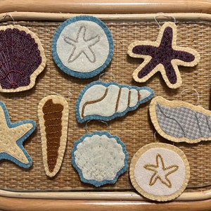 May include: A collection of felt seashell ornaments in various shapes and colors, including starfish, sand dollars, and conch shells. Each ornament has a light blue border and is embellished with beads and embroidery. The ornaments are displayed on a woven surface.