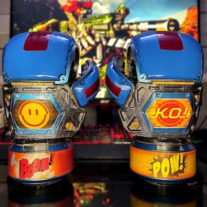 May include: Pair of blue and red boxing gloves with comic book style text that says "BOOM!" and "POW!". The gloves have a smiley face and "K.O.!" on the sides. The background shows a blurred image of a video game.