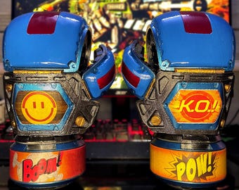 Puede incluir: Un par de guantes de boxeo azules y rojos con texto estilo cómic que dice "BOOM!" y "POW!". Los guantes tienen una carita sonriente y "K.O.!" en los lados. El fondo muestra una imagen borrosa de un videojuego.