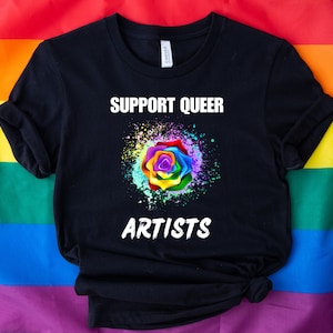 May include: Black t-shirt with the text "Support Queer Artists" in white lettering. A rainbow rose with a white splatter effect is centered on the shirt.