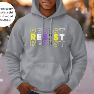 May include: A gray hoodie with the word "RESIST" printed in white and yellow letters. The letters are stacked on top of each other, with the "S" in the word "RESIST" being purple. The hoodie is being worn by a person with a dark complexion. The text "$10 from every sweatshirt sold will be donated to the ACLU" is in the top left corner of the image.
