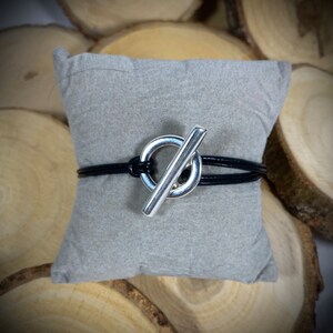 May include: A black leather bracelet with a silver toggle clasp. The clasp is a circle with a bar across it.