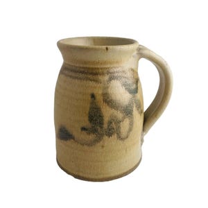 May include: A handmade ceramic mug with a beige base color and a dark brown abstract design. The mug has a rounded body, a sturdy handle, and a slightly flared rim. The mug is suitable for hot or cold beverages.