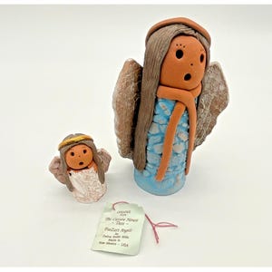 May include: Two ceramic angel figurines. The larger angel has brown hair, a blue dress, and outstretched wings. The smaller angel has a gold halo and white dress. Both have surprised expressions. A tag reads "PooZart Angels".